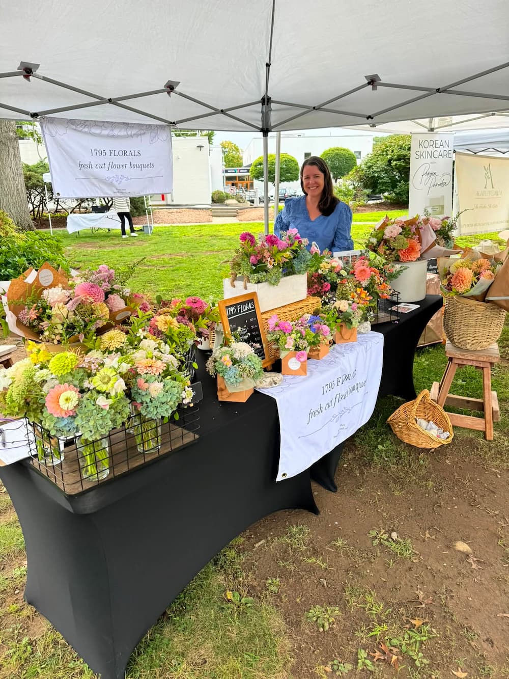 An outdoor flower market stall under a white canopy tent, displaying an assortment of colorful flower bouquets, potted plants, and baskets on a black tablecloth-covered table. A woman in a blue shirt stands behind the display, with a banner reading '1795 Florals fresh cut flower bouquets' visible. Market display at the Wilton Farmers' Market with Stacey from 1795 Florals