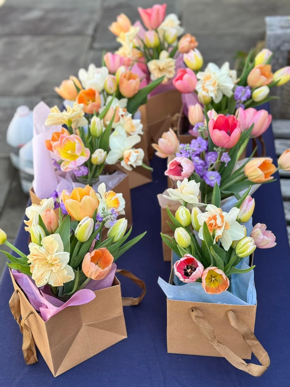 Several small flower bouquets arranged in brown paper gift bags on a dark blue table outdoors. The bouquets contain tulips, daisies, and purple flowers in pastel colors including pink, orange, yellow, and white. Tulip Bouquets from 1795 Florals.