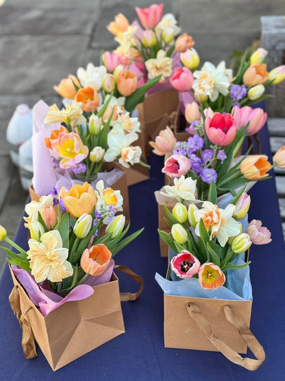 Several small flower bouquets arranged in brown paper gift bags on a dark blue table outdoors. The bouquets contain tulips, daisies, and purple flowers in pastel colors including pink, orange, yellow, and white. Tulip Bouquets from 1795 Florals.