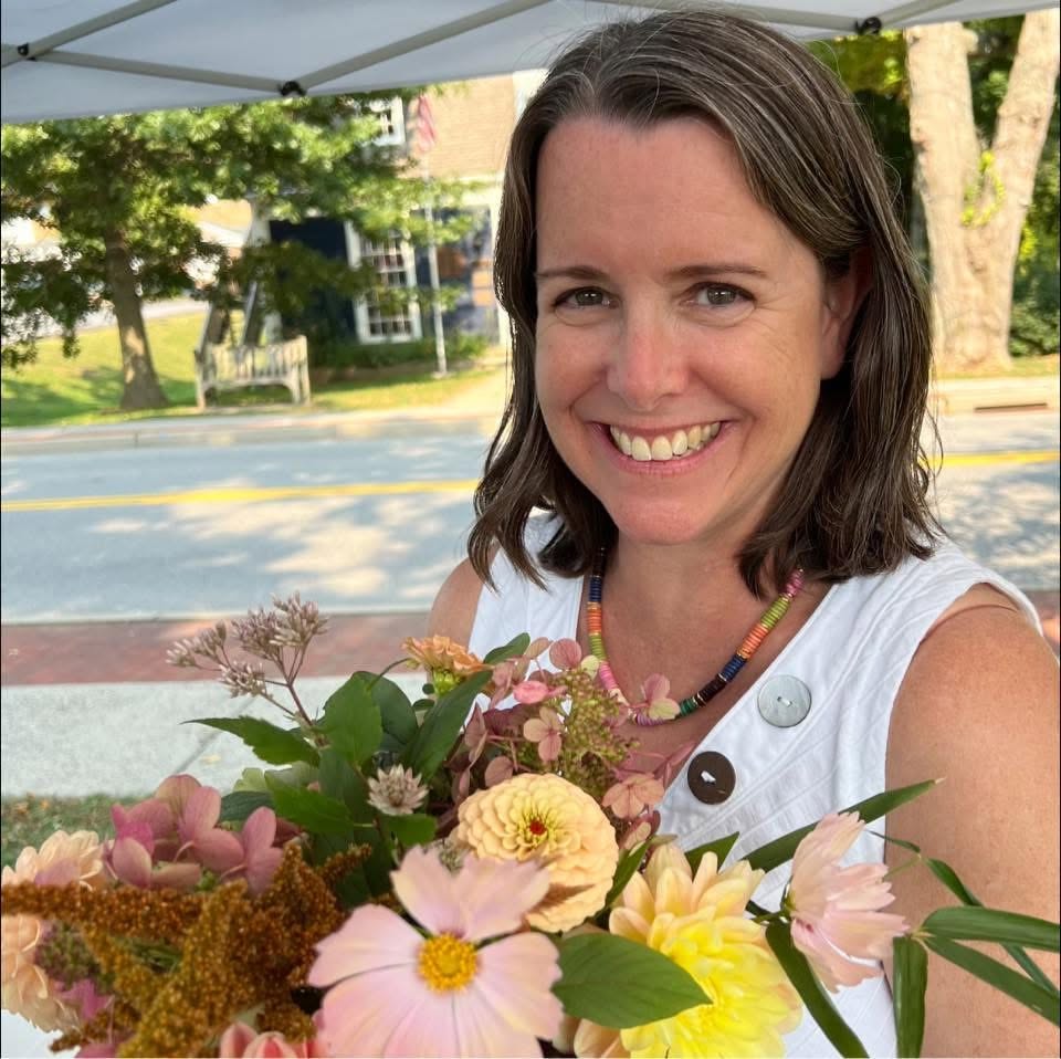 A smiling woman with shoulder-length brown hair stands outdoors under a white canopy tent, holding a colorful bouquet of pink, yellow, and peach flowers. She is wearing a white sleeveless top and a colorful beaded necklace, with a street and trees visible in the background. Stacey from 1795 Florals in Wilton, CT