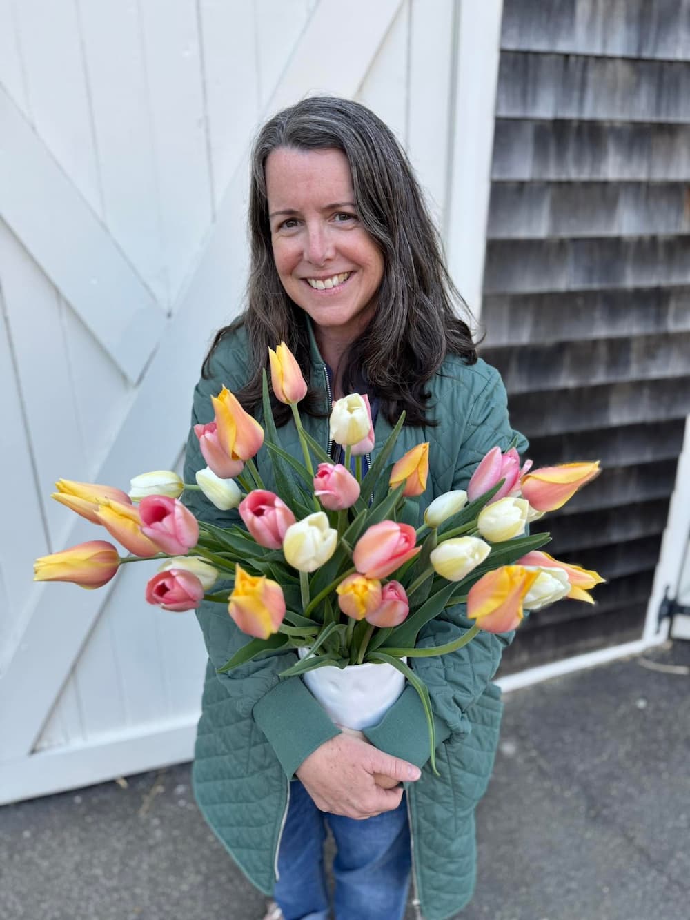 A smiling woman with long brown hair stands outdoors holding a large bouquet of tulips in pink, yellow, and white. She is wearing a quilted green jacket and blue jeans, positioned in front of a white wooden barn door and a gray shingled wall. Stacey Sapper from 1795 Florals, located in Wilton, CT.